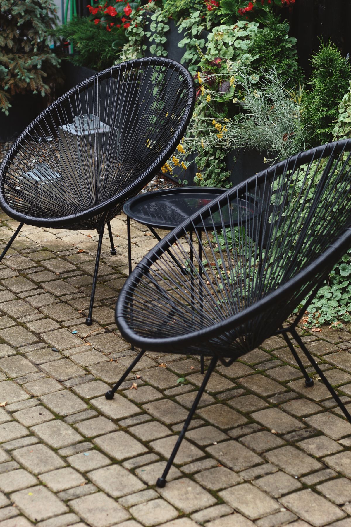 Two empty black lounge chairs tucked beside a leafy patio garden